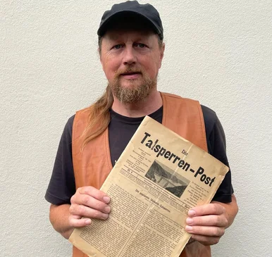 Der Referent Eckhart Böhm mit der Zeitschrift „Die Talsperren-Post“ Eckhart Böhm, ein Mann mittleren Alters mit Kappe und Zopf, hält die historische Zeitschrift Die Talsperren-Post in der Hand
