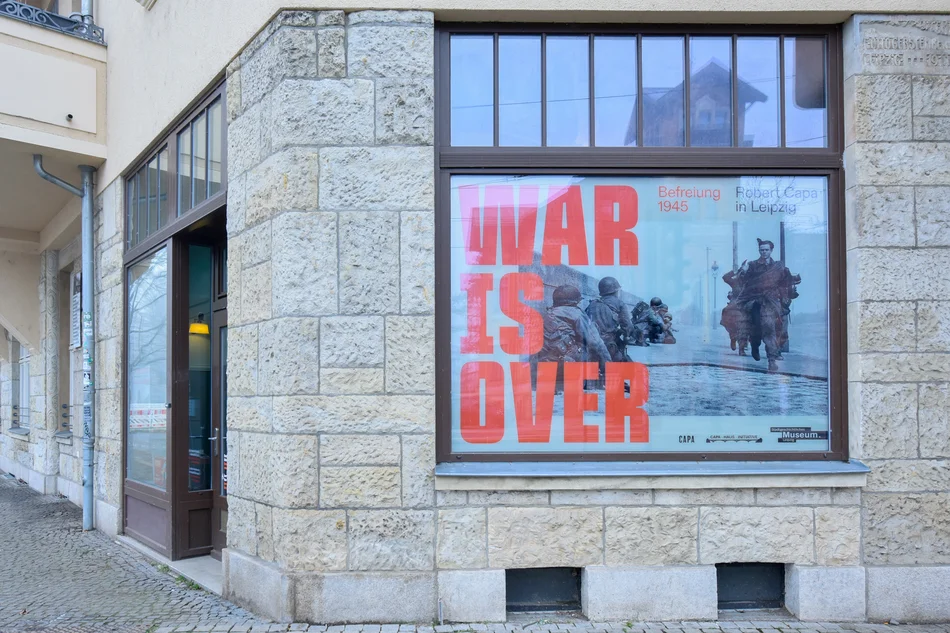 Ausstellung „War Is Over“ im Capa-Haus, Leipzig Das Erdgeschossfenster eines Eckhauses wird von einem Ausstellungsplakat eingenommen, darauf in großen roten Lettern der Schriftzug "WAR IS OVER" vor einem Foto von Robert Capa im April 1945 zur Befreiung von Leipzig