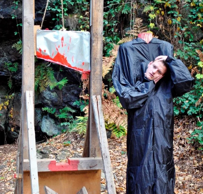 Neben der Attrappe einer mit roter Farbe beschmierten Guillotine steht eine mannshohe Puppe die ihren Kopf in den Händen hält