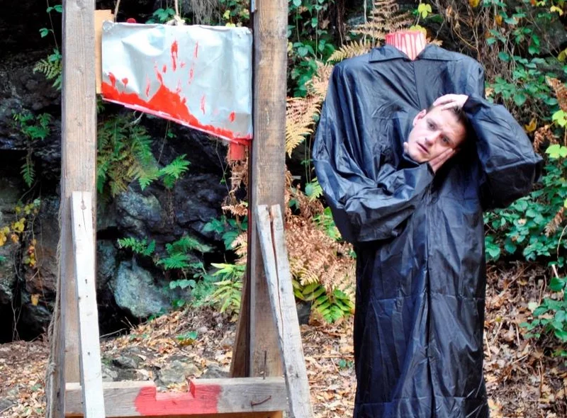 Neben der Attrappe einer mit roter Farbe beschmierten Guillotine steht eine mannshohe Puppe die ihren Kopf in den Händen hält