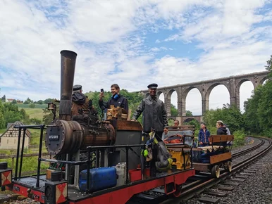 Die Dampfdraisine kommt zu Gast ins Chemnitztal. Die Dampfdraisine mit einem Anhänger steht auf Gleisen unterhalb einer Bogenbrücke. Der Lokführer schaut freundlich.