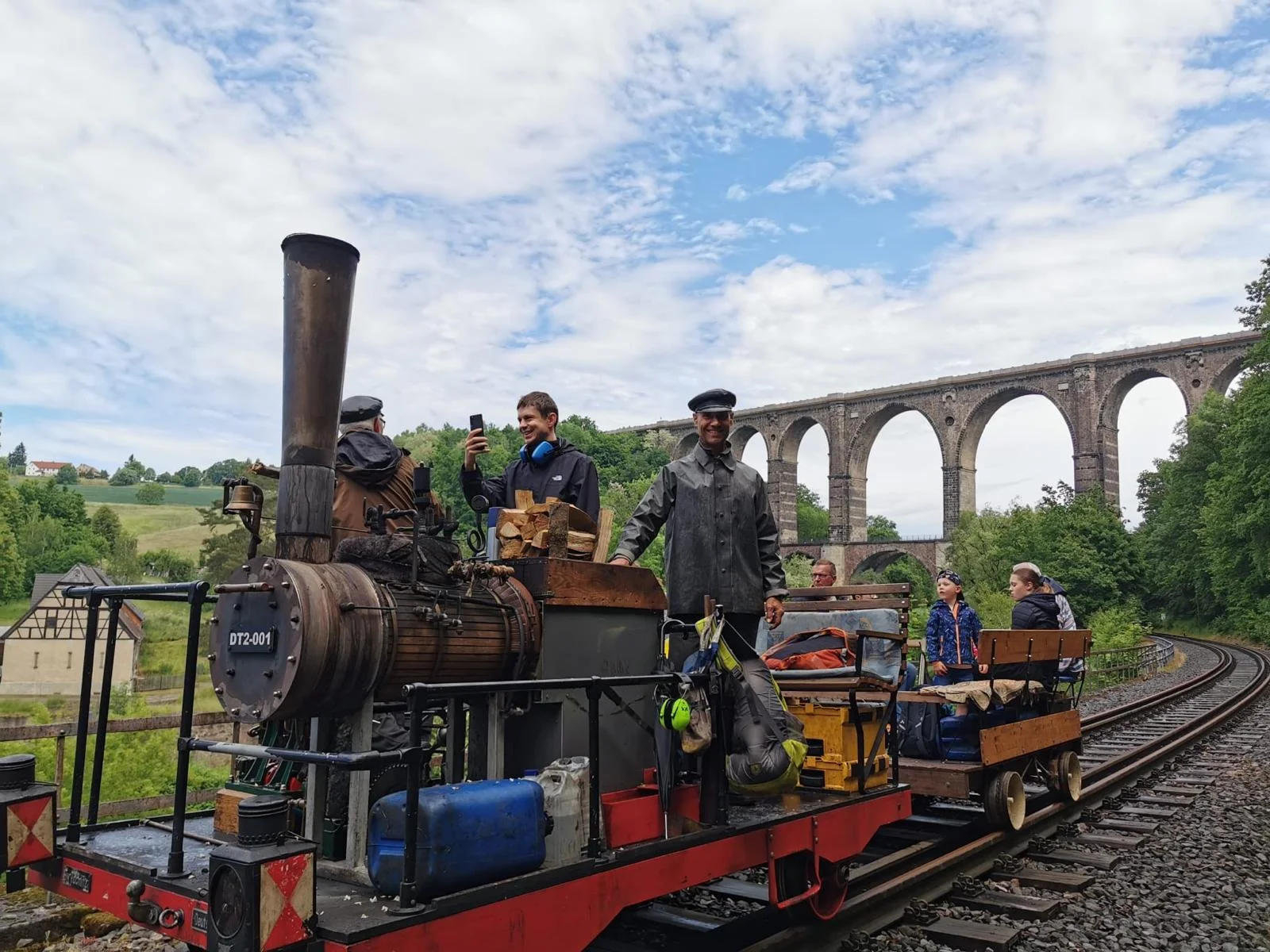 Die Dampfdraisine kommt zu Gast ins Chemnitztal. Die Dampfdraisine mit einem Anhänger steht auf Gleisen unterhalb einer Bogenbrücke. Der Lokführer schaut freundlich.