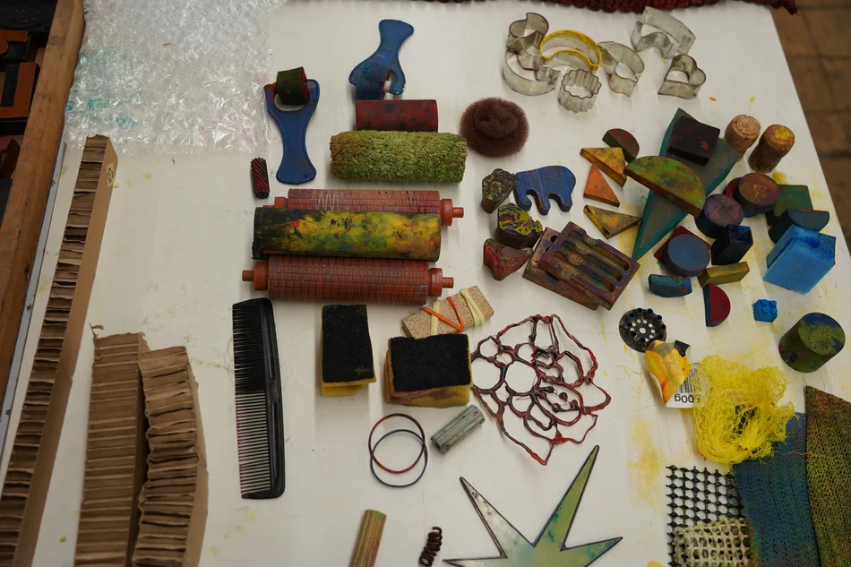Auf einer Arbeitsfläche sind verschiedenste Gegenstände zum Drucken angeordnet – von einem Kamm, einem Netz und Gummibändern über diverse Keksausstecher bis hin zu Holzblöcken und Druckerrollen.