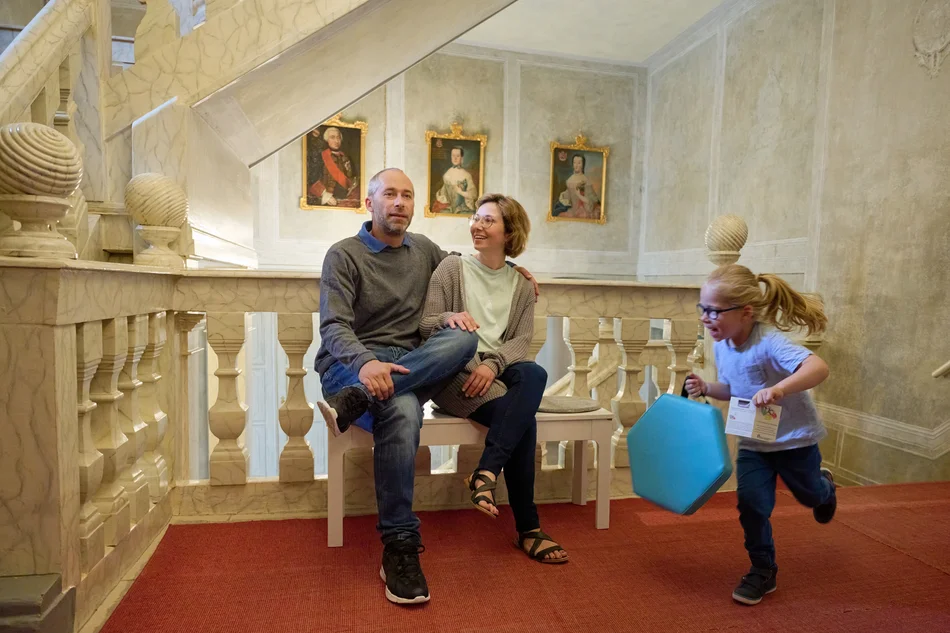 Zwei Erwachsene sitzen in einem historischen Treppenhaus. Ein Kind mit Sitzkissen rennt fröhlich an den beiden vorbei.