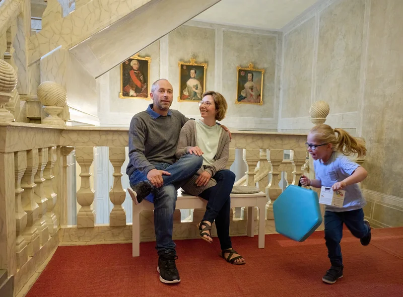 Zwei Erwachsene sitzen in einem historischen Treppenhaus. Ein Kind mit Sitzkissen rennt fröhlich an den beiden vorbei.
