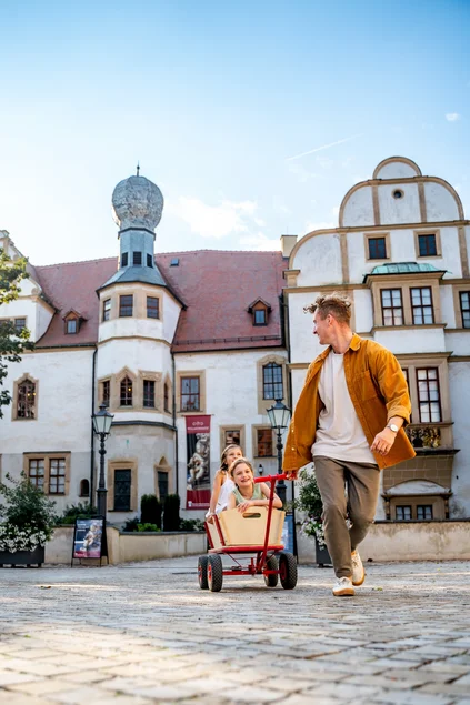 Zwei lachende Kinder im Handwagen, die von einer Person vor Schlosskulisse über den Hof gezogen werden