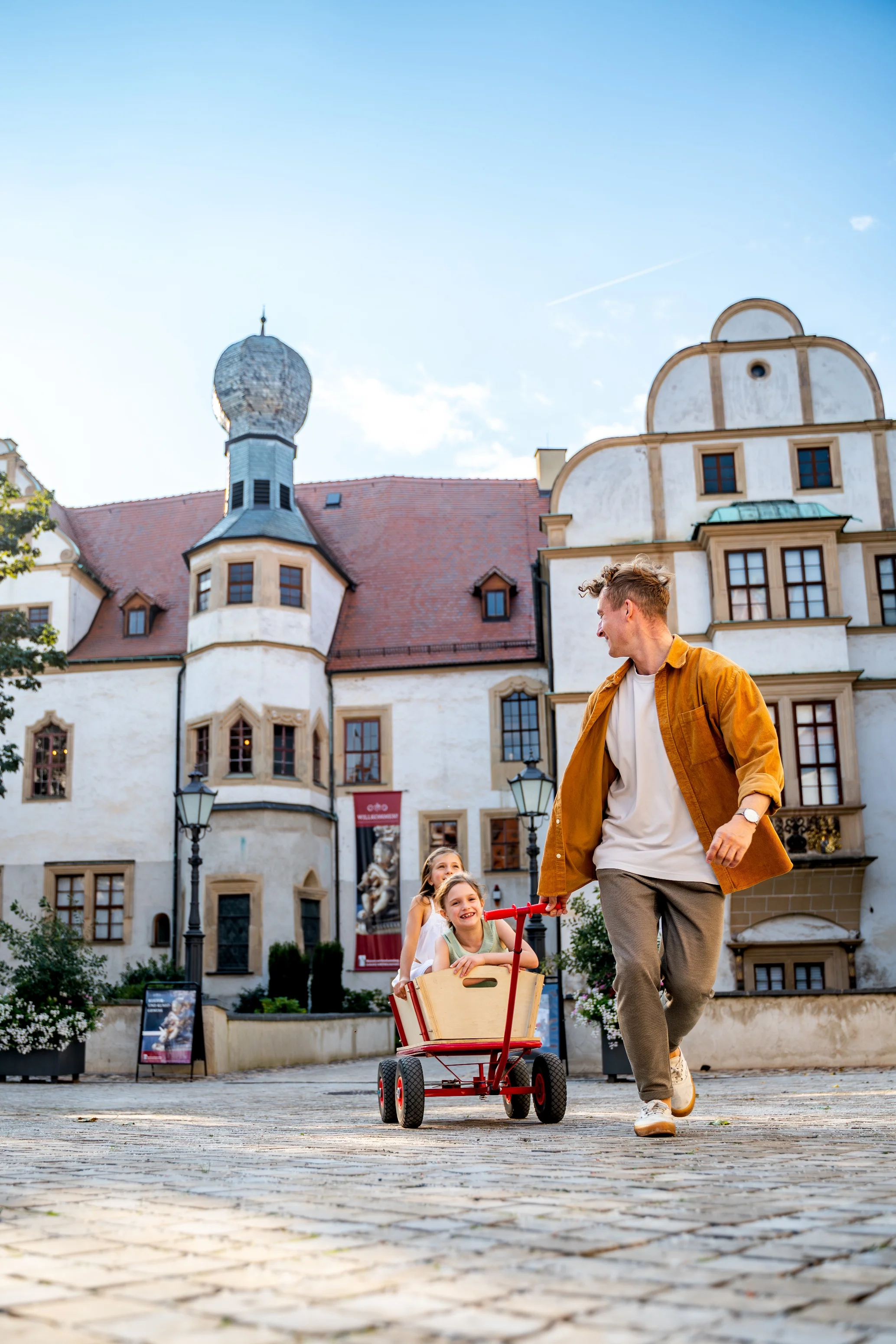 Zwei lachende Kinder im Handwagen, die von einer Person vor Schlosskulisse über den Hof gezogen werden