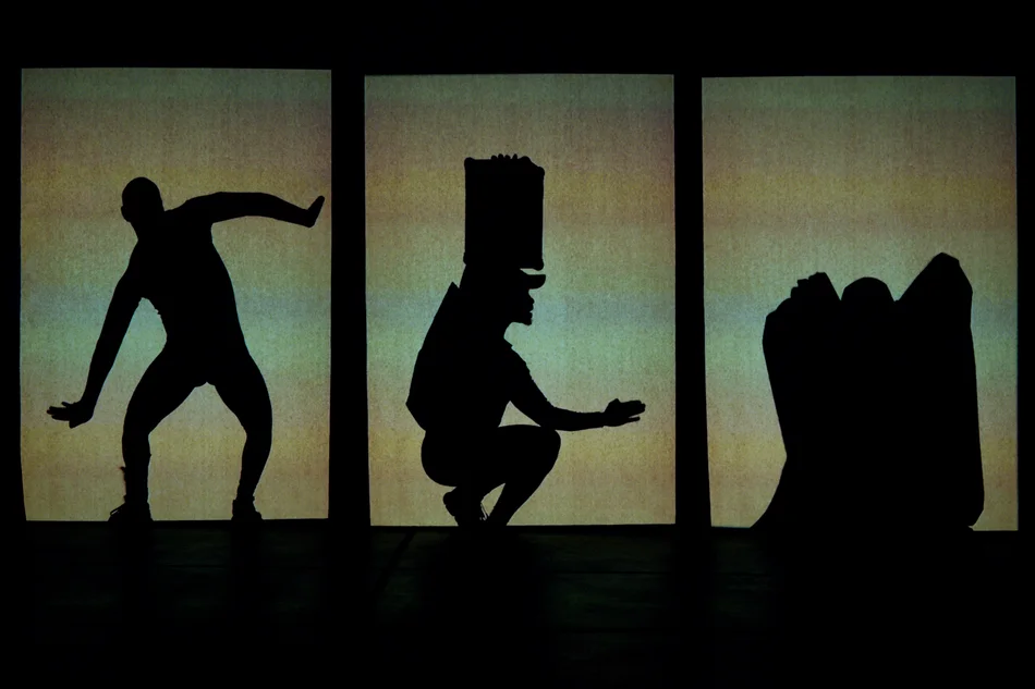 Eine Projektionsleinwand, die vertikal in drei Segmente unterteilt ist. In jedem der Felder ist die schwarze Silhouette eines Tänzers zu erkennen – in unterschiedlichen Posen und mit unterschiedlichen Requisiten und Kleidungsstücken