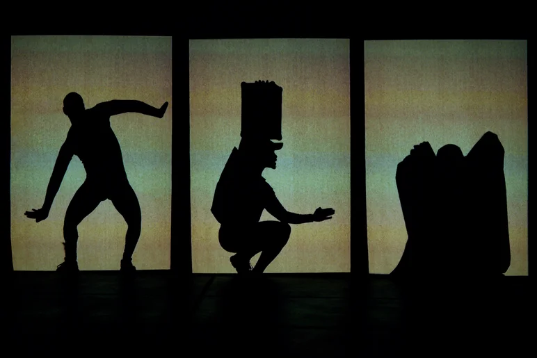 Eine Projektionsleinwand, die vertikal in drei Segmente unterteilt ist. In jedem der Felder ist die schwarze Silhouette eines Tänzers zu erkennen – in unterschiedlichen Posen und mit unterschiedlichen Requisiten und Kleidungsstücken