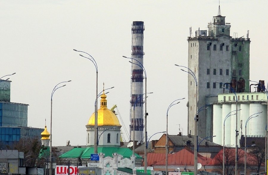 Detail einer urban-industriellen Landschaft mit den goldenen Zwiebeltürmen einer russisch-orthodoxen Kirche im Vordergrund und einem Schornstein und Industriebauten im Hintergrund. Der Himmel ist grau.