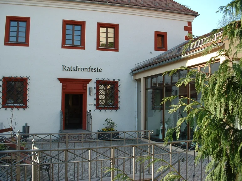 Weiß getünchte Fassade eines mehrstöckigen historischen Gebäudes mit in weinroter Farbe gefassten Kassettenfenstern und einem weinrot gefassten schmalen Portal, welches den Eingang ins Museum darstellt. Über dem Portal ist der schwarze Schriftzug „Ratsfronfeste“ angebracht.