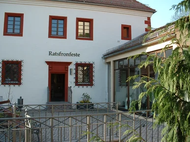 Weiß getünchte Fassade eines mehrstöckigen historischen Gebäudes mit in weinroter Farbe gefassten Kassettenfenstern und einem weinrot gefassten schmalen Portal, welches den Eingang ins Museum darstellt. Über dem Portal ist der schwarze Schriftzug „Ratsfronfeste“ angebracht.