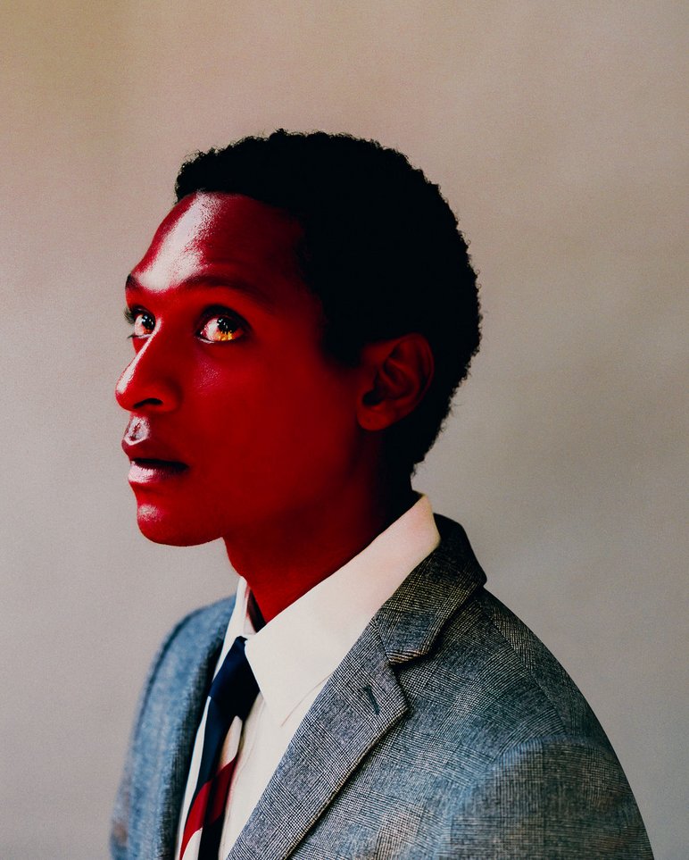 Diese Fotografie von Aaron Ricketts zeigt Kopf- und Schulterpartie eines jungen afrikanischen oder afroamerikanischen Mannes im Seitenprofil. Seine Haut ist glänzend-rot gefärbt. Gekleidet in Anzug und Krawatte, richtet er seinen Blick nach schräg oben.