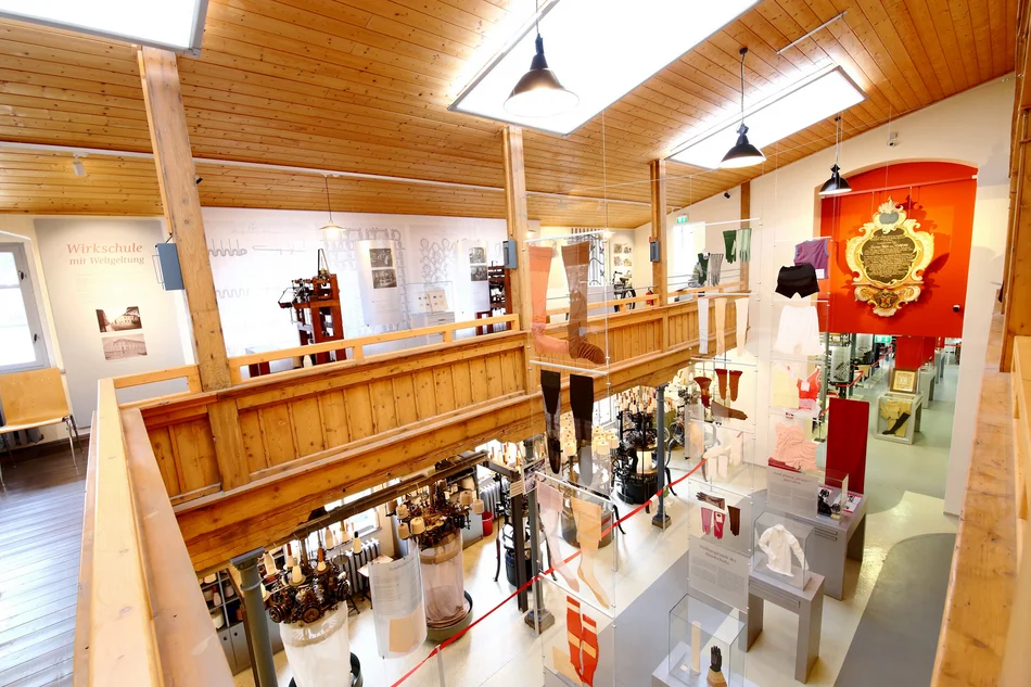 Das Foto gibt einen Blick in die Dauerausstellung. Aufgenommen von der Empore im ersten Obergeschoss, auf der unter anderem historische Handwirkstühle gezeigt werden, schweift der Blick nach unten in den ehemaligen Maschinensaal der alten Wirkerei, wo unter anderem Rundkulierwirkmaschinen, mehrere in der Esche-Wirkerei hergestellte Strümpfe und Handschuhe sowie weitere Maschinen und Textilien ausgestellt sind.