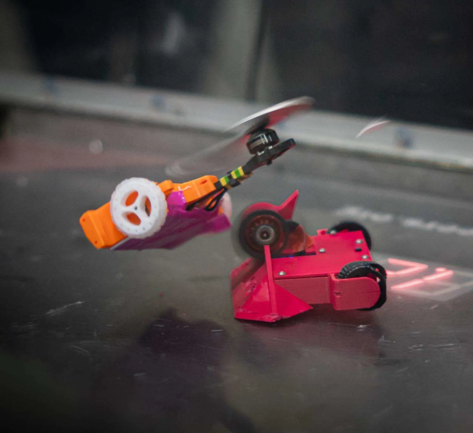 Robot Combat Zwei kleine Roboter im Duell: Der rote fährt auf Rädern, der orangefarbene scheint wie eine Mini-Drohne zu fliegen.