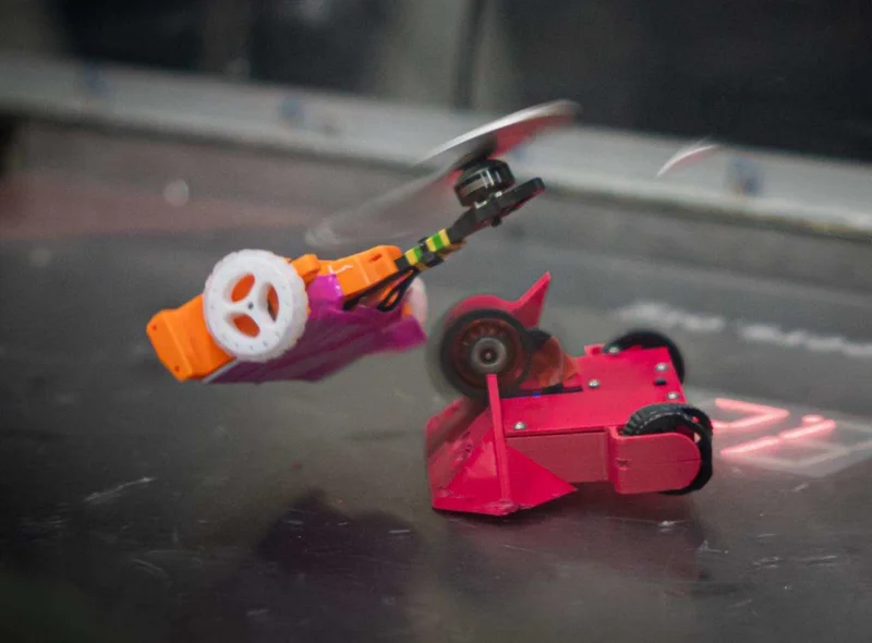 Zwei kleine Roboter im Duell: Der rote fährt auf Rädern, der orangefarbene scheint wie eine Mini-Drohne zu fliegen.