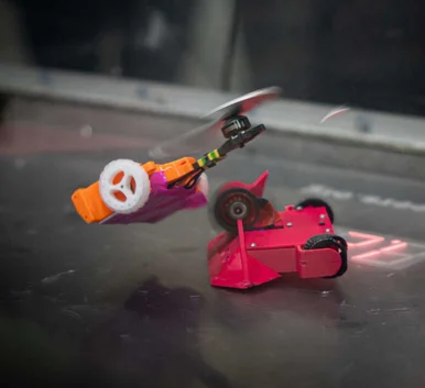 Robot Combat Zwei kleine Roboter im Duell: Der rote fährt auf Rädern, der orangefarbene scheint wie eine Mini-Drohne zu fliegen.