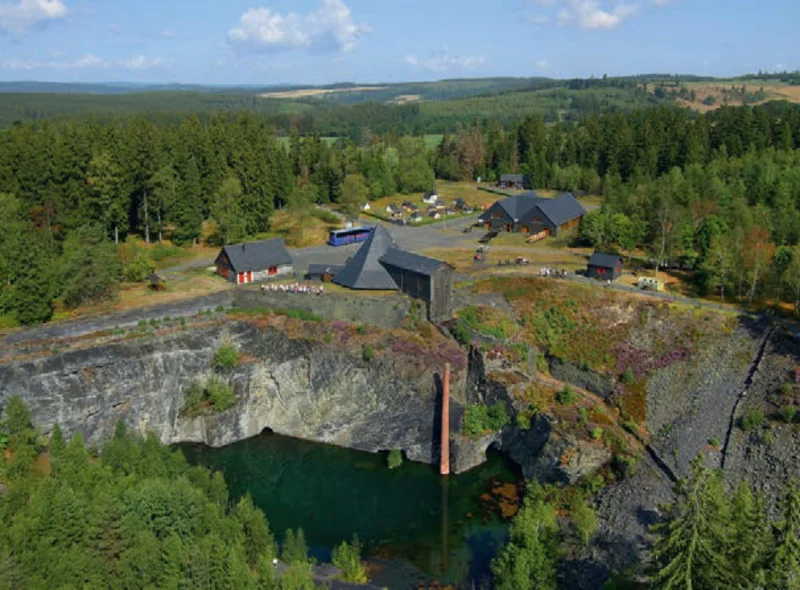 Vogelperspektive auf einen mit Wasser gefüllten Steinbruch, umrandet von Wald, daneben einige kleine Gebäude