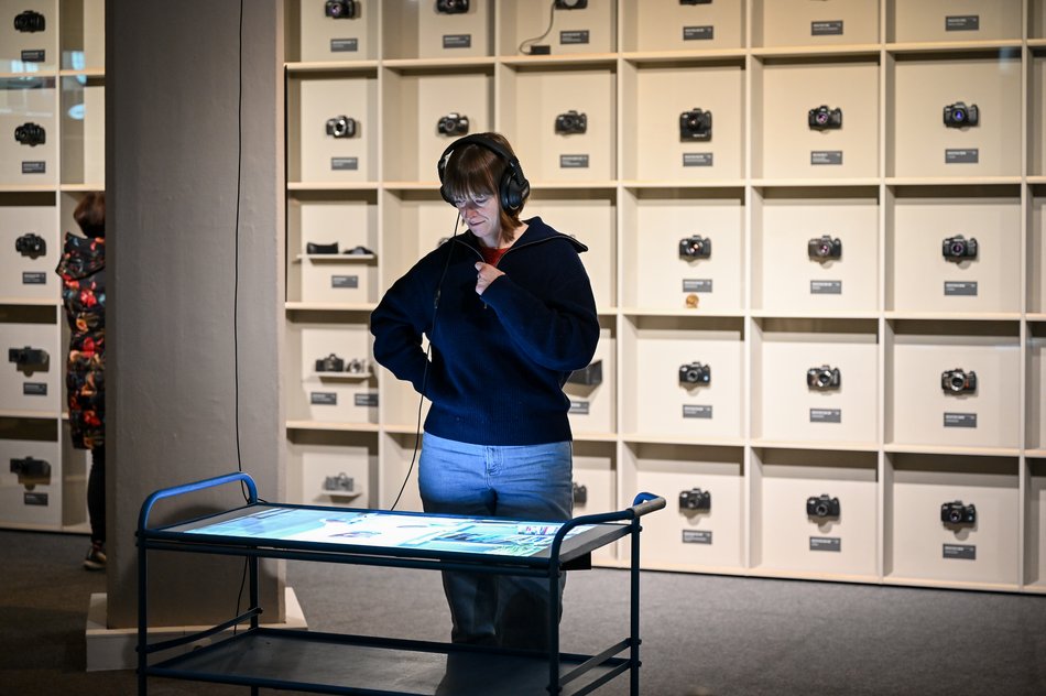 Eine Frau mit Kopfhörern steht an einer Medienstation und betrachtet beim Hören die Bilder vor ihr. Die Wand im Hintergrund wird vollständig von einem Regal eingenommen, in jedem der quadratischen Regalfächer ist ein historischer Fotoapparat ausgestellt.