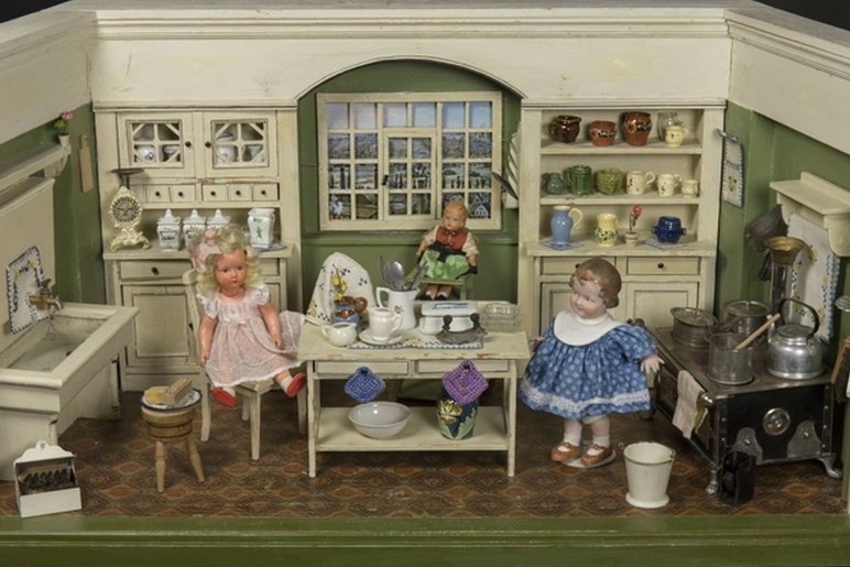 Das Foto zeigt eine Puppenküche aus der Zeit um 1920. Um den Küchentisch herum sitzen drei Puppen auf Stühlen: zwei weibliche Erwachsene und ein Kleinkind. An der Wand hinter ihnen befinden sich Regale mit Küchenutensilien, rechterhand ein eiserner Herd, linkerhand ein großes Spülbecken.