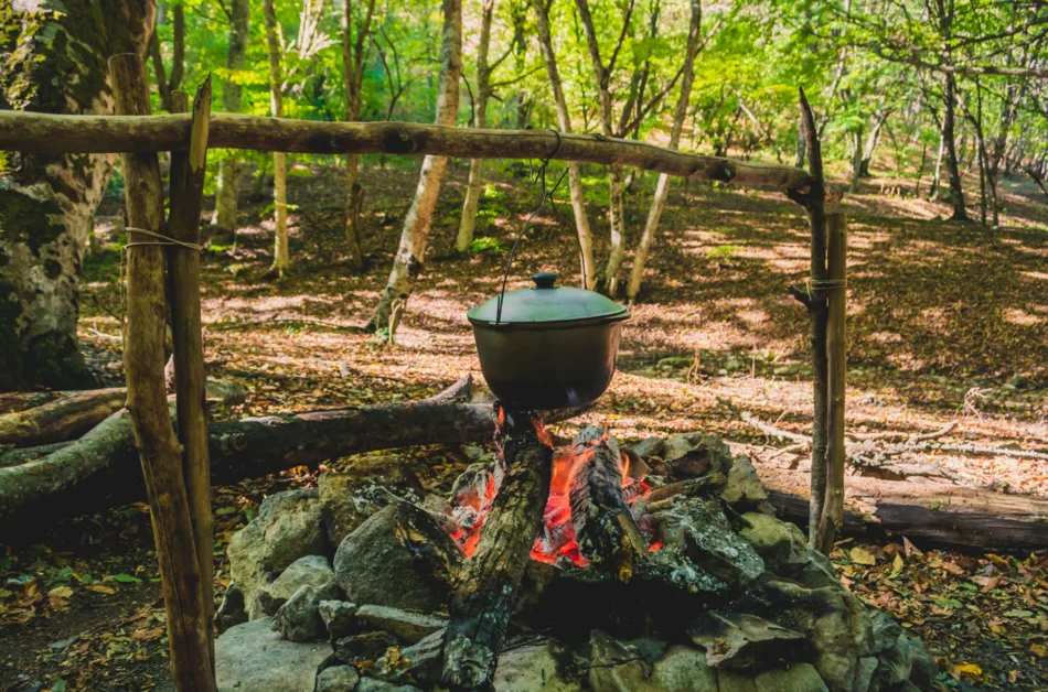 Über einer Feuerstelle im Wald hängt ein gusseisener Kochtopf mit Deckel.
