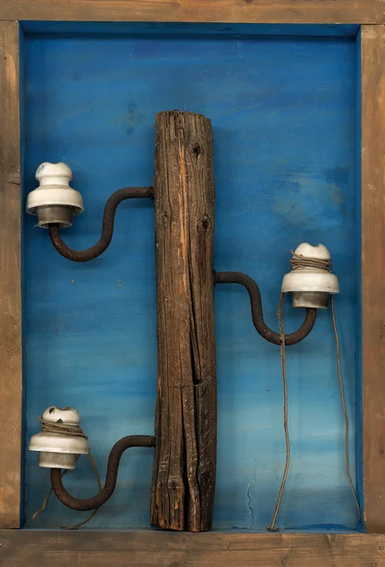 Der obere Teil eines historischen Strommastes mit Holzpfahl, drei Porzellan-Isolatoren und herunterhängenden Kabeln, assembliert in einem Holzrahmen mit himmelblau bemaltem Hintergrund