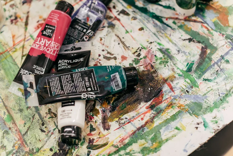 Das Foto zeigt vier halbvolle Acrylfarbtuben auf einer mit bunten Pinselstrichen und Klecksen bedeckten weißen Fläche.