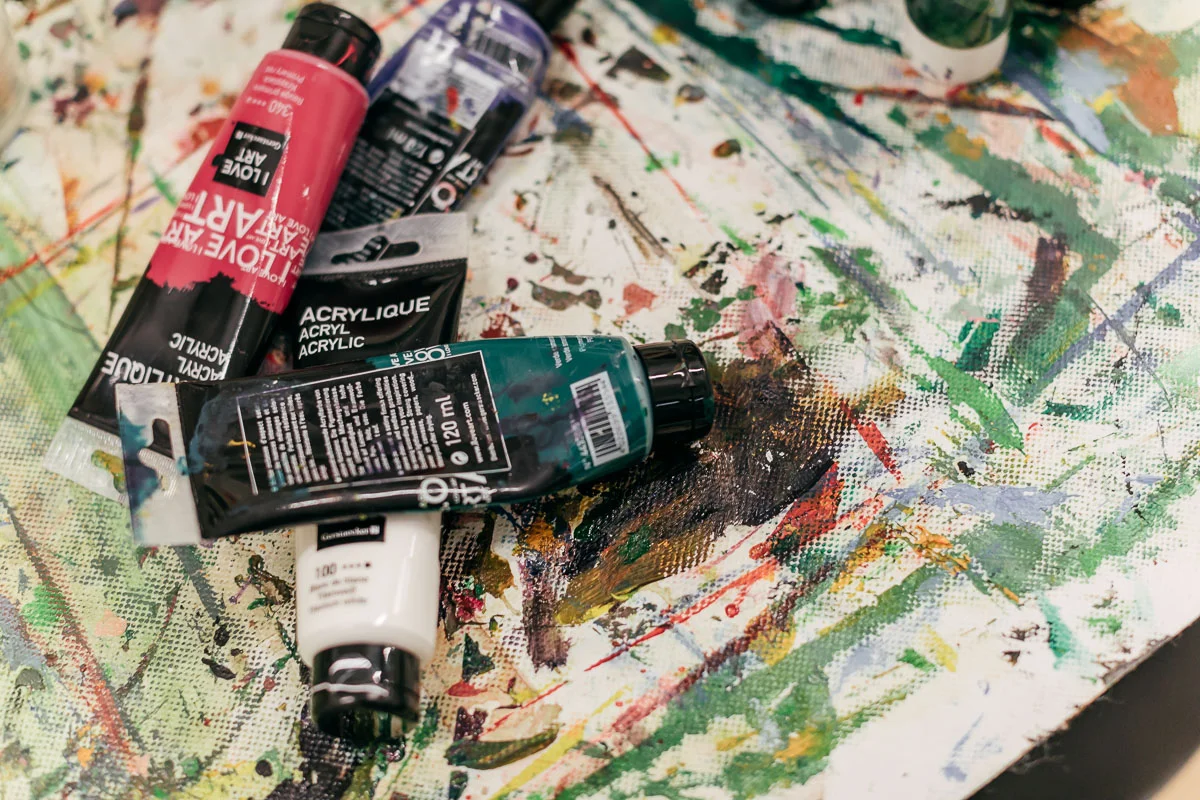 Das Foto zeigt vier halbvolle Acrylfarbtuben auf einer mit bunten Pinselstrichen und Klecksen bedeckten weißen Fläche.