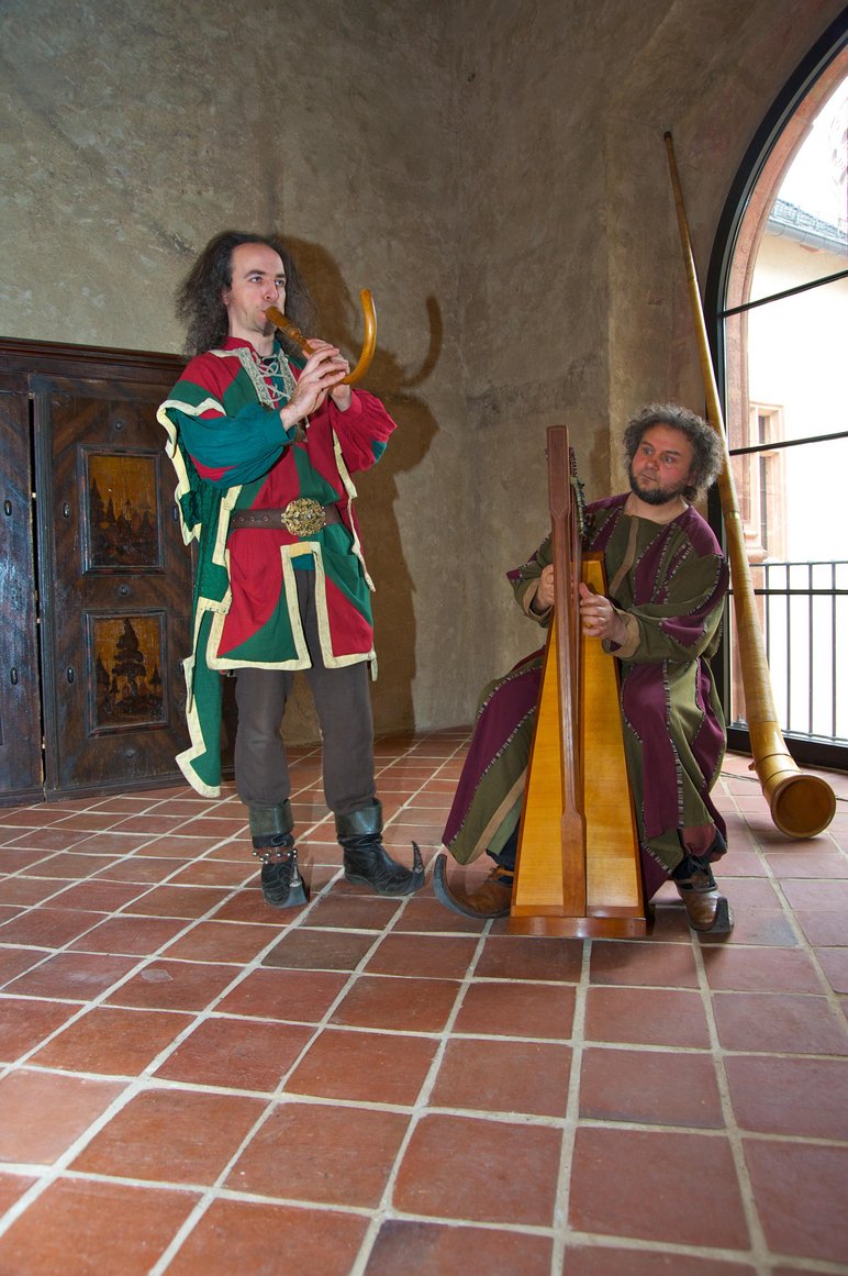 Zwei in Mittelalter-Kostüme gekleidete Männer spielen in einem Raum von Burg Mildenstein auf historischen Instrumenten.