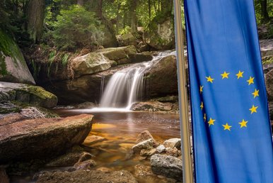 Eine EU-Fahne vor einem idyllischen Gewässer mit kleinem Wasserfall