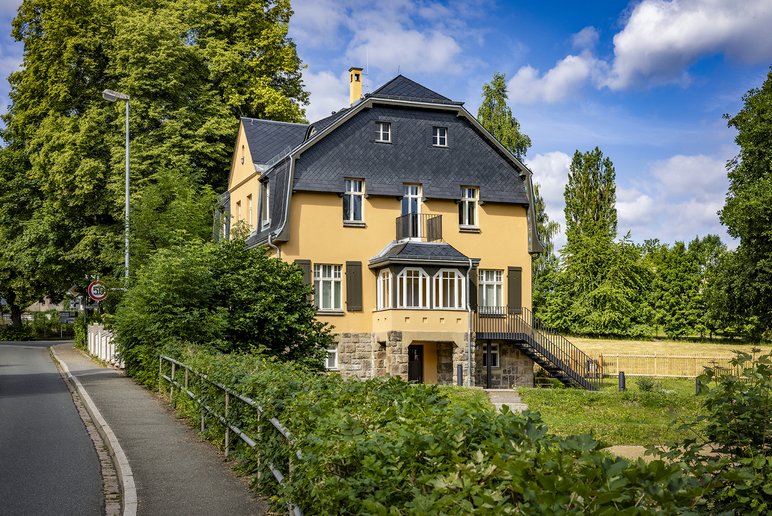 Sommerliche Außenansicht einer Villa aus dem frühen 20. Jahrhundert mit sandfarbener Fassade und schiefergedecktem Dachfirst