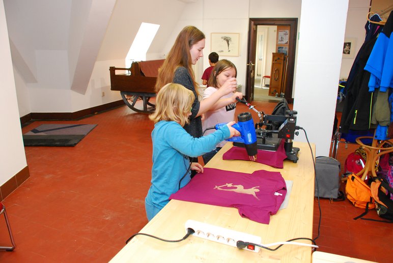 Das Foto zeigt Kinder, die unter Anleitung einer Erwachsenen T-Shirts im Siebdruckverfahren bedrucken.