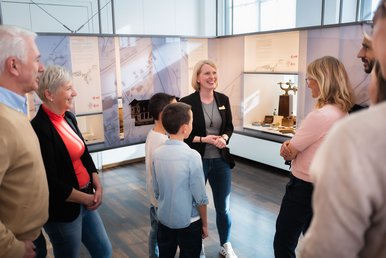 In einem mit Vitrinen bestückten Ausstellungsraum spricht eine Museumspädagogin zu einer im Halbkreis um sie herum stehenden Personengruppe.