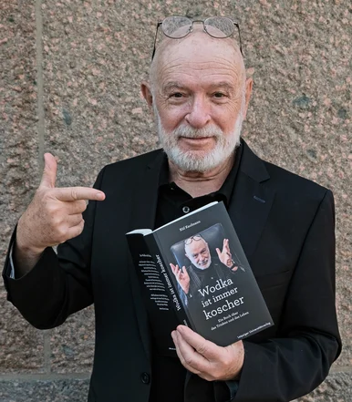 Porträtfoto eines grauhaarigen Mannes mit Brille und Bart, der mit einer Hand das Buch „Wodka ist immer koscher“ hält und mit der anderen Hand darauf zeigt