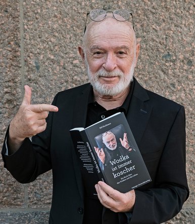 Küf Kaufmann Porträtfoto eines grauhaarigen Mannes mit Brille und Bart, der mit einer Hand das Buch „Wodka ist immer koscher“ hält und mit der anderen Hand darauf zeigt