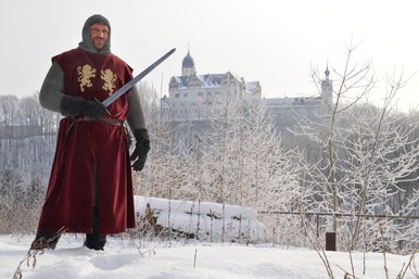Foto eines als mittelalterlicher Ritter verkleideten Mannes mit Schwert in winterlicher Kulisse, im Hintergrund Schloss Rochsburg