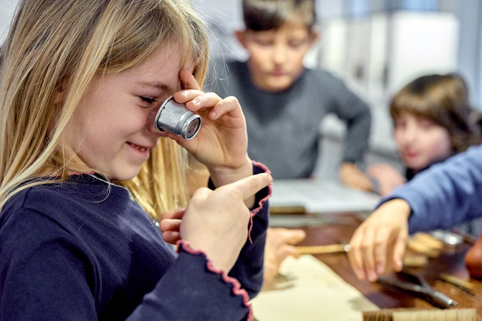 Das Foto zeigt ein Mädchen mit Uhrmacherlupe und zwei weitere Kinder im Hintergrund, die im Deutschen Uhrenmuseum Glashütte die Zeit entdecken.
