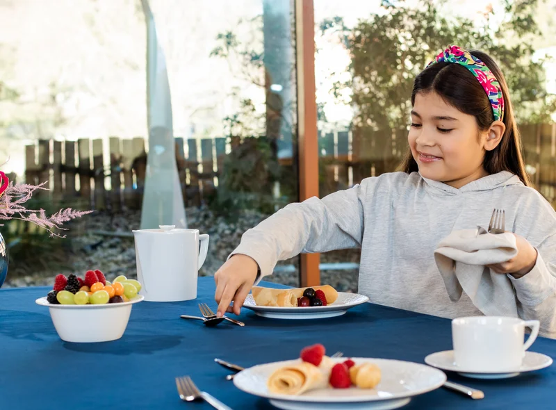 Ein Mädchen an einem mit weißem Porzellan gedeckten Tisch hebt mit Besteck Obstsalat auf ihren Teller.