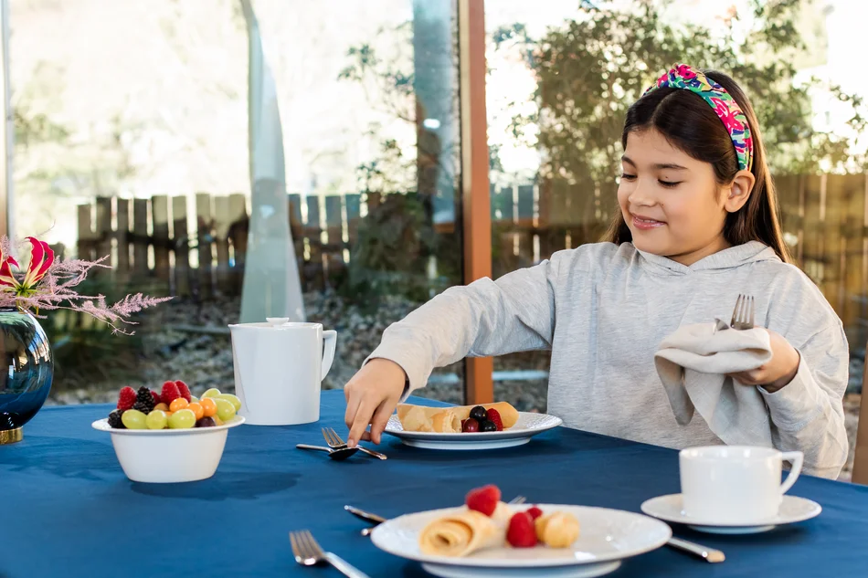 Ein Mädchen an einem mit weißem Porzellan gedeckten Tisch hebt mit Besteck Obstsalat auf ihren Teller.