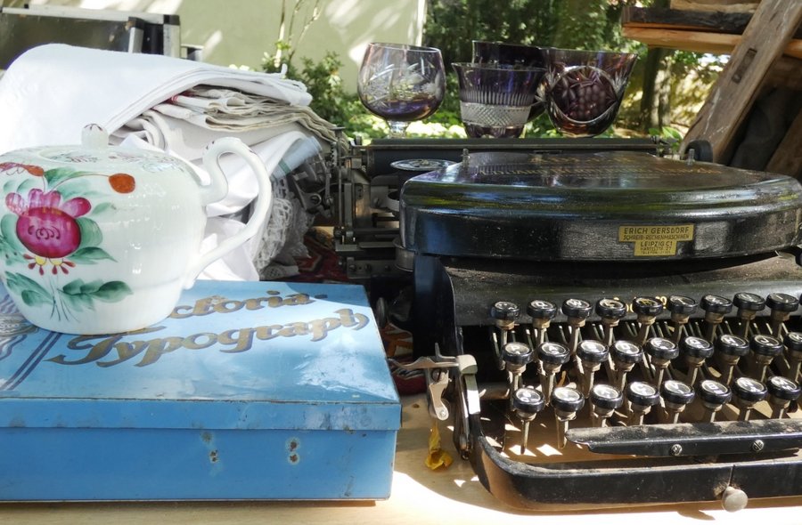 Nahaufnahme eines Flohmarkt-Tisches, auf dem neben einer historischen Schreibmaschine ein mit Blumendekor verziertes Teekännchen, eine hellblauer Blechdose, ein kleiner Stapel weißer Tischdecken sowie einige geschliffene Weingläser stehen
