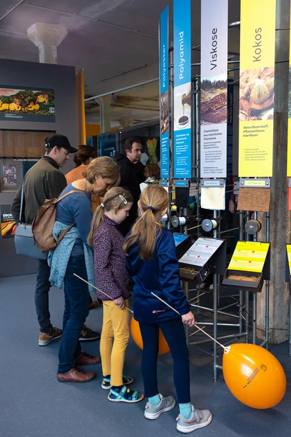Turm der Tuche An einem kreisförmig angeordneten Ausstellungselement, an dem Text- und Bildtafeln angebracht sind, stehen mehrere Besucherinnen und Besucher.