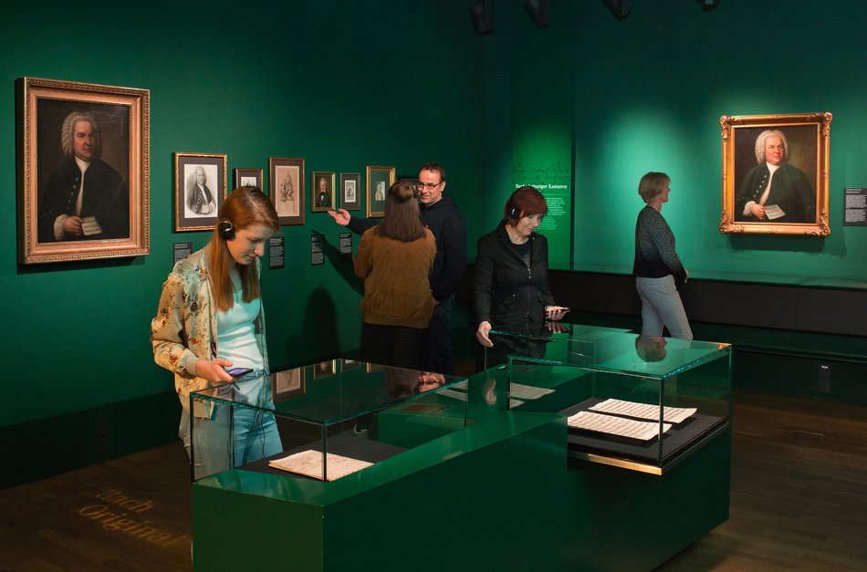 Das Foto zeigt einen Blick in das Herzstück des Bach-Museums Leipzig: die Schatzkammer. Zu sehen sind fünf Besucherinnen und Besucher, zwei davon mit Audioguides, Objekte in Tischvitrinen betrachtend.