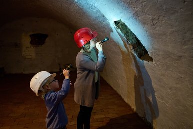 Abwärts. Taschenlampenführung mit Schatzsuche Eine Frau und ein kleiner Junge, beide mit Helmen ausgestattet, stehen in einem historischen Kellergang und leuchten mit Taschenlampen auf eine Stelle an der Wand.
