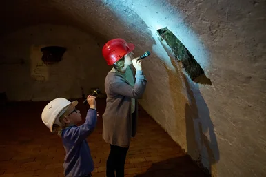 Eine Frau und ein kleiner Junge, beide mit Helmen ausgestattet, stehen in einem historischen Kellergang und leuchten mit Taschenlampen auf eine Stelle an der Wand.