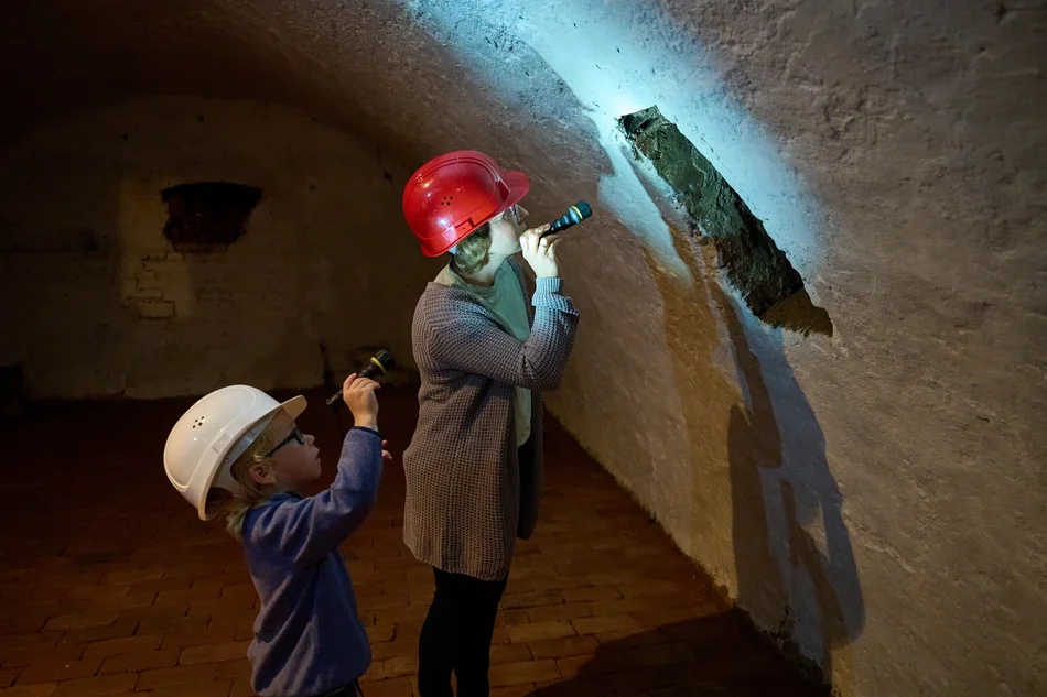 Eine Frau und ein kleiner Junge, beide mit Helmen ausgestattet, stehen in einem historischen Kellergang und leuchten mit Taschenlampen auf eine Stelle an der Wand.