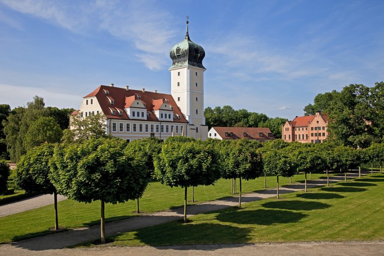 In einer sommerlich-grünen Parkanlage mit Rasenflächen und beschnittenen Bäumchen steht ein Schloss mit Zwiebeltürmchen.