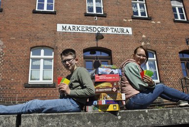 Brettspielabend auf dem Museumsbahnhof Ein Junge und ein Mädchen sitzen auf der Bahnsteigkante des Museumsbahnhofs Markersdorf-Taura, zwischen ihnen ein Stapel Brettspiele.