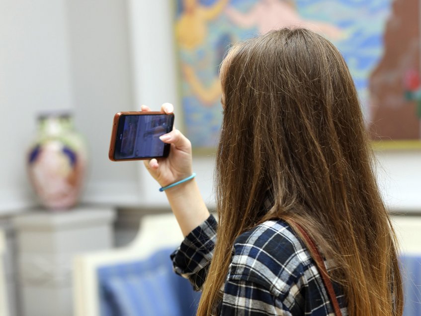 Von schräg hinten ist eine langhaarige junge Frau in einer Kunstausstellung zu sehen, die ihre Smartphone-Kamera auf ein Exponat richtet.