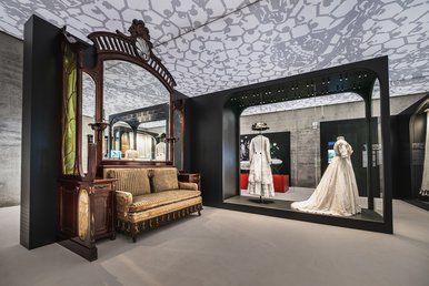 Blick in die Ausstellung der Fabrik der Fäden Mitten im Raum steht eine edel wirkende Vitrine mit zwei Gewändern in weißer Plauener Spitze. Daneben steht vor einem großen in verziertem Holz gefassten Spiegel ein Sofa im Biedermeier-Stil.