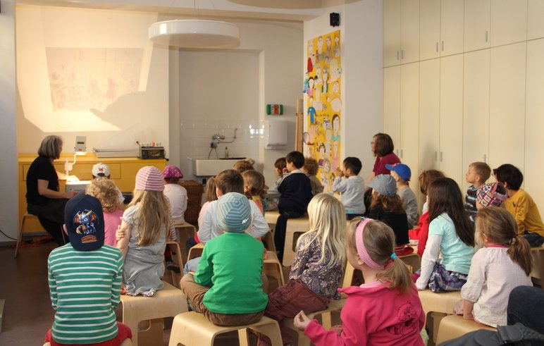Das Foto zeigt, wie eine Kindergruppe bei einer Lesung mit Bildern und Musik zuhört.